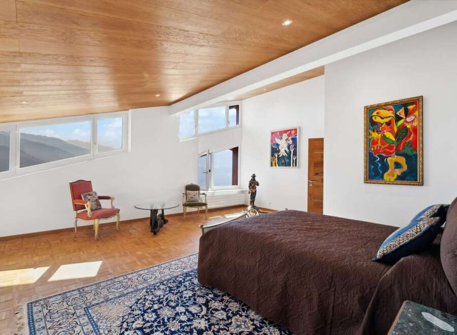 This luxurious bedroom boasts stunning mountain views through its uniquely shaped windows, complemented by a wooden ceiling and hardwood floors.