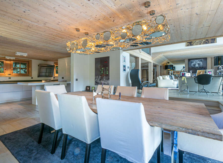 This stunning chalet boasts a spacious dining area with a rustic wooden table and modern white chairs, illuminated by a unique circular chandelier.