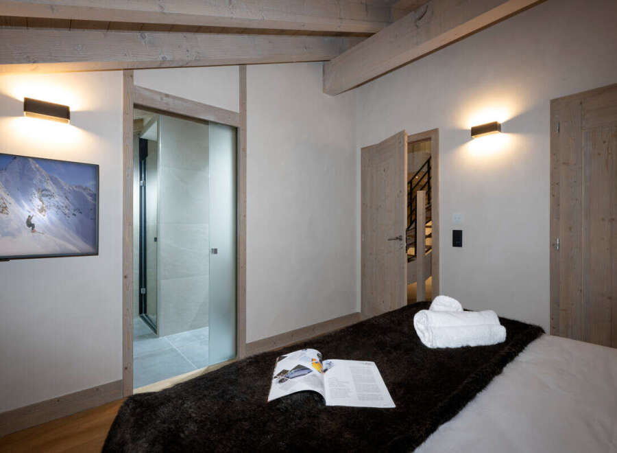 This luxurious bedroom boasts a stunning mountain view from its large TV, an ensuite bathroom with glass doors, and a cozy bed with a faux fur throw.
