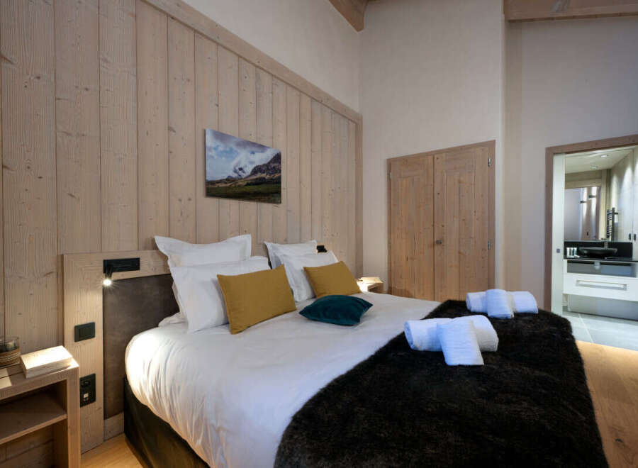 This luxurious bedroom boasts a king-size bed dressed in crisp white linens and plush dark fur throw, complemented by a stunning mountain landscape painting.