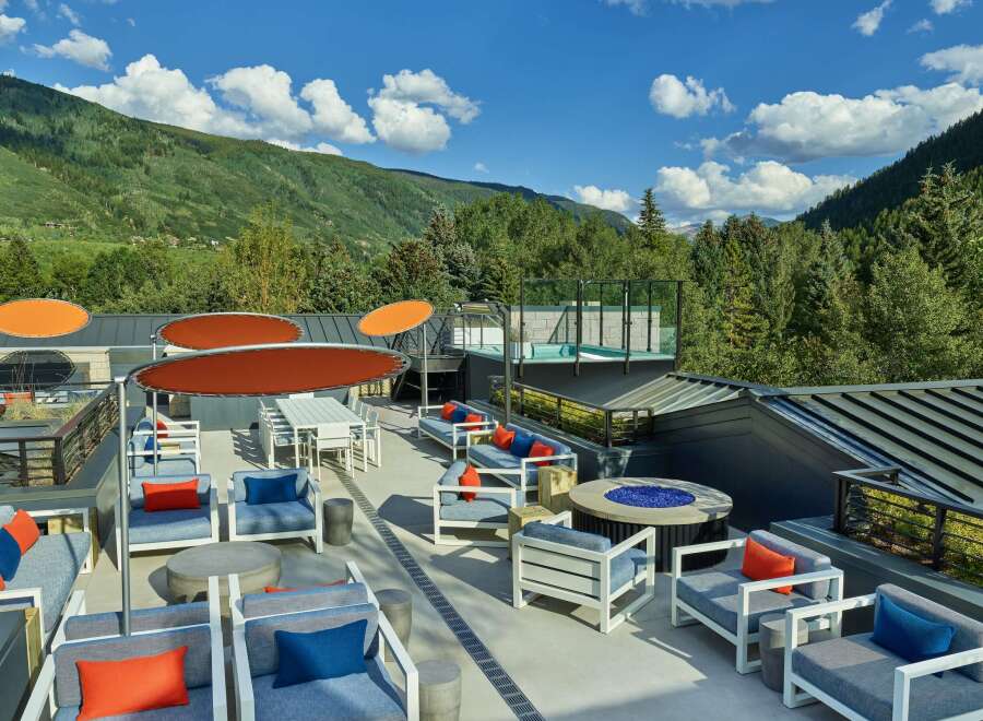 This rooftop patio boasts stunning mountain views, comfortable seating areas with vibrant orange and blue pillows, and a fire pit for cozy gatherings.