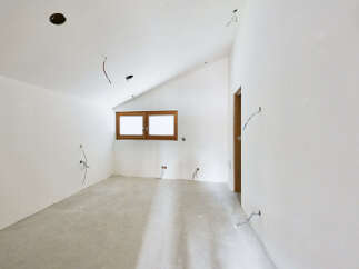 This unfinished room features a sloped ceiling with recessed lighting, a large window providing ample natural light, and exposed wiring indicating ongoing renovations.