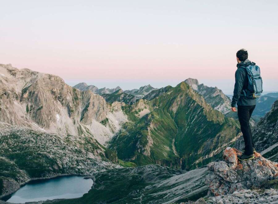 A hiker stands on a rocky outcrop, gazing out at a breathtaking vista of rolling green and grey mountains.