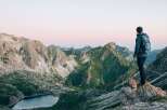 A hiker stands on a rocky outcrop, gazing out at a breathtaking vista of rolling green and grey mountains.