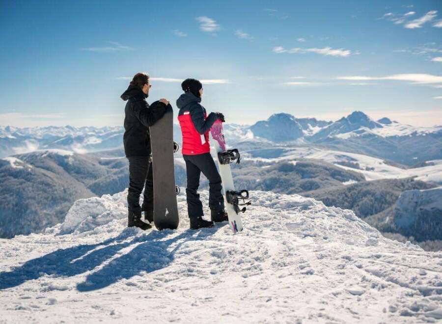 Two snowboarders stand atop a snowy mountain peak, gazing out at a breathtaking panoramic view of the snow-covered mountain range.