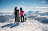 Two snowboarders stand atop a snowy mountain peak, gazing out at a breathtaking panoramic view of the snow-covered mountain range.