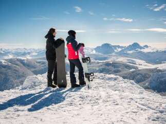 Two snowboarders stand atop a snowy mountain peak, gazing out at a breathtaking panoramic view of the snow-covered mountain range.