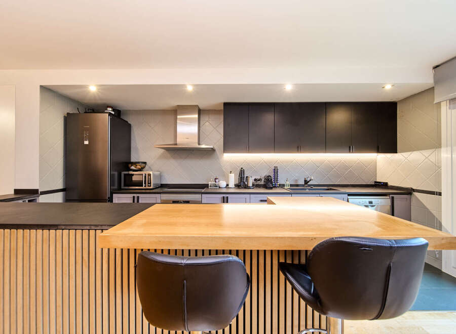 Modern kitchen with sleek dark cabinets, light gray backsplash, and a light wood island with bar stools.