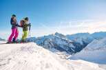 Skiers enjoy breathtaking panoramic views atop a snow-covered mountain, with pristine slopes and a stunning backdrop of snow-capped peaks.
