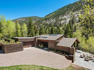 This stunning mountain home boasts a spacious, gravel driveway leading to a rustic-chic log cabin with a modern flat roof extension.