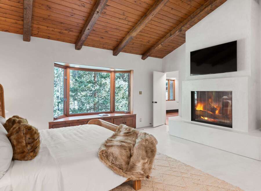 This stunning bedroom boasts a cozy fireplace, a large bay window overlooking a snowy landscape, and a wood-beamed ceiling.
