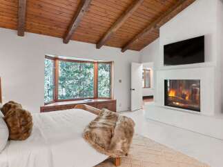 This stunning bedroom boasts a cozy fireplace, a large bay window overlooking a snowy landscape, and a wood-beamed ceiling.