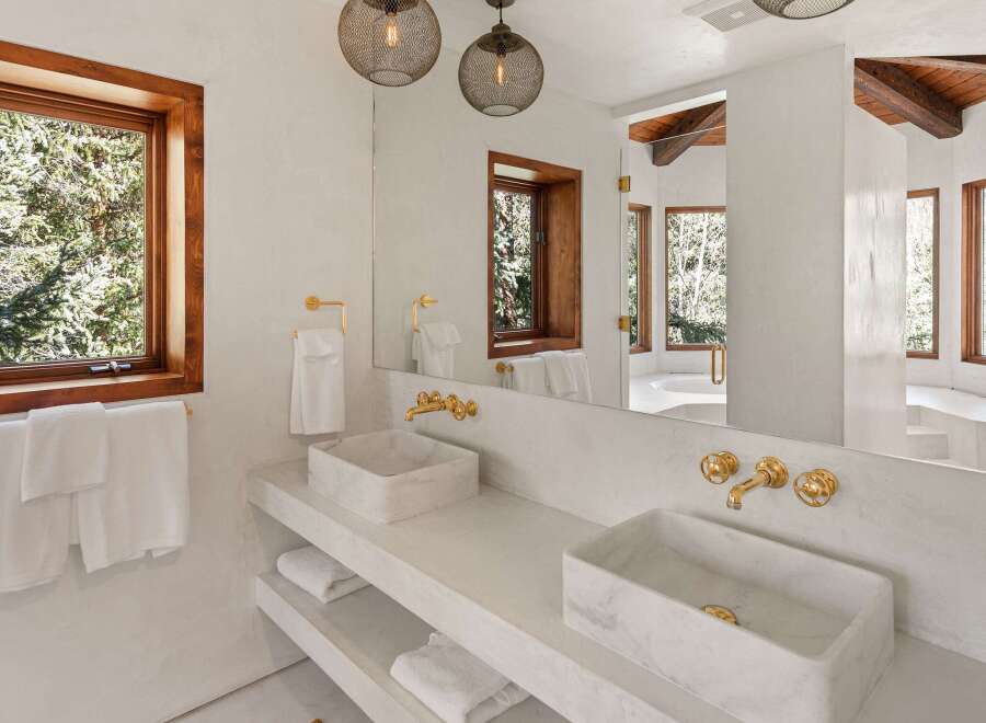 This luxurious bathroom boasts a stunning double vanity with marble sinks and gold fixtures, complemented by large windows offering breathtaking views of the surrounding trees.