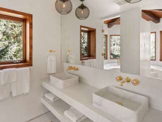 This luxurious bathroom boasts a stunning double vanity with marble sinks and gold fixtures, complemented by large windows offering breathtaking views of the surrounding trees.