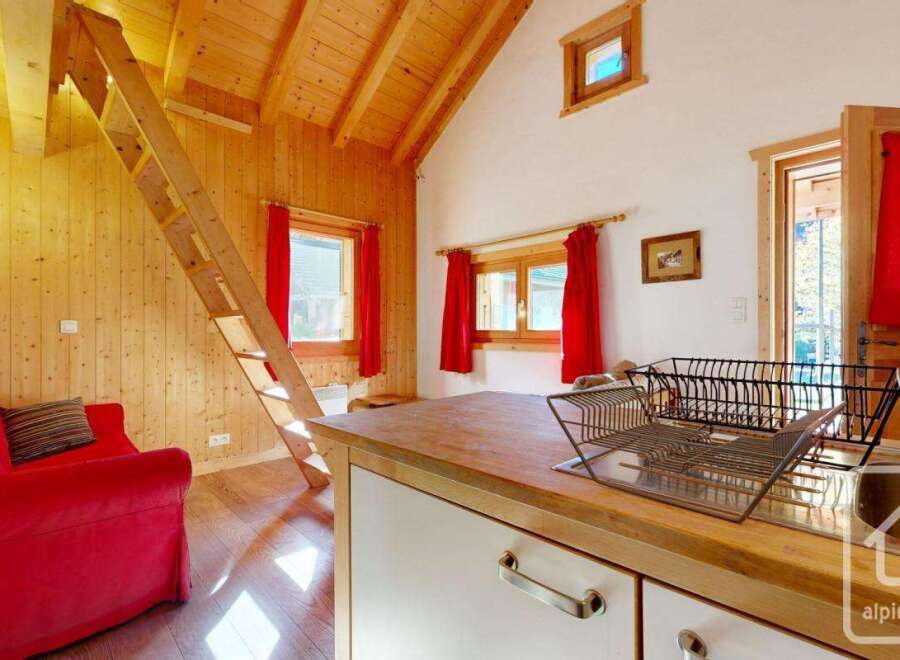 Cozy chalet apartment with wooden interior, featuring a red sofa, a wooden staircase leading to a loft, and a small kitchen with wooden countertops.
