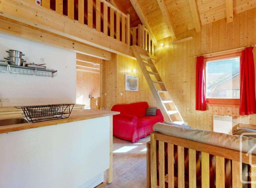 Cozy chalet apartment featuring a well-equipped kitchenette, a comfortable red sofa, and a charming wooden loft bedroom accessible by a ladder.