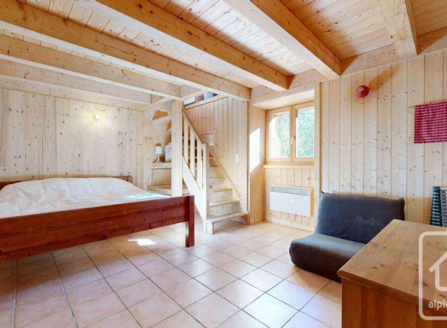 Cozy chalet bedroom with wooden walls and ceiling, featuring a double bed, a small sofa, and a wooden staircase leading to an upper level.