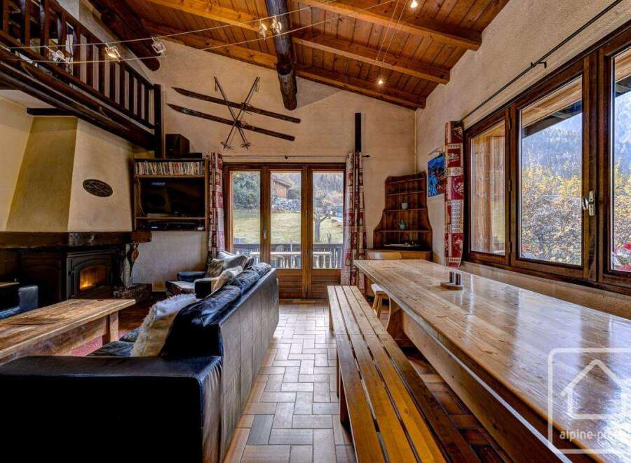 This charming chalet boasts a spacious open-plan living area with a large wooden dining table perfect for gatherings, a cozy fireplace for chilly evenings, and ample natural light from large windows showcasing stunning mountain views.