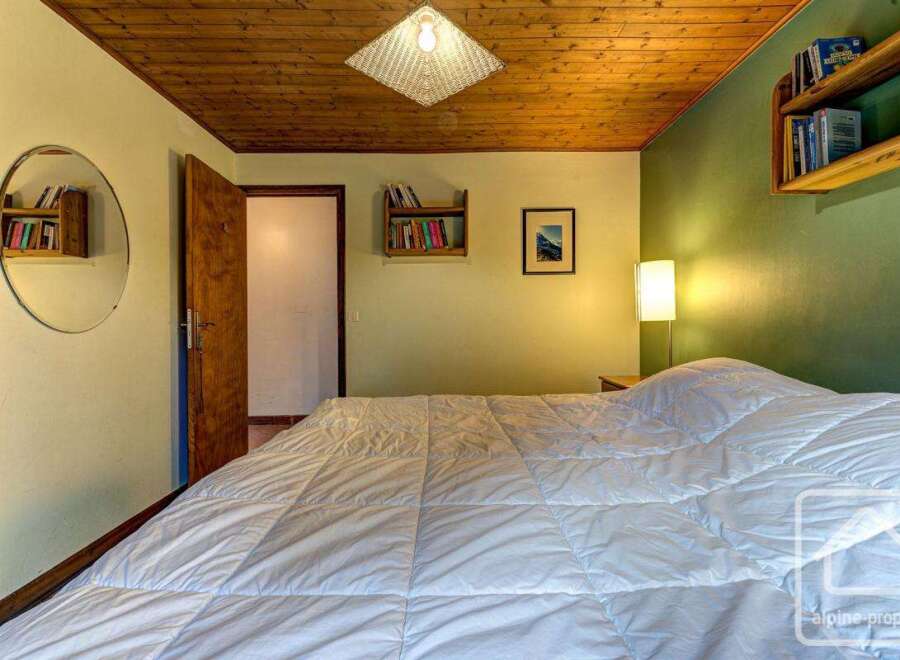 Cozy bedroom with wooden ceiling and walls, featuring a comfortable double bed with a white duvet, built-in bookshelves, and a round mirror.
