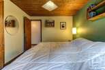 Cozy bedroom with wooden ceiling and walls, featuring a comfortable double bed with a white duvet, built-in bookshelves, and a round mirror.