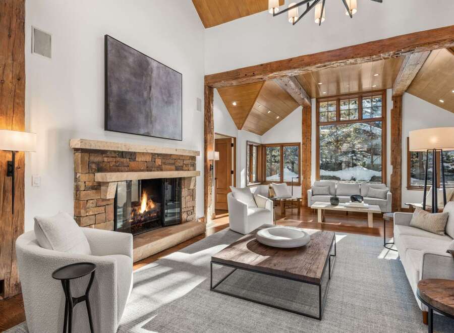 This stunning living room boasts a cozy stone fireplace, large windows offering breathtaking views, and a high ceiling with exposed wooden beams.