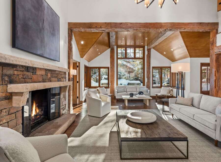 This stunning living room boasts a rustic-chic design with exposed wooden beams, a stone fireplace, and large windows offering breathtaking views of a snow-covered landscape.