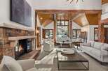 This stunning living room boasts a rustic-chic design with exposed wooden beams, a stone fireplace, and large windows offering breathtaking views of a snow-covered landscape.