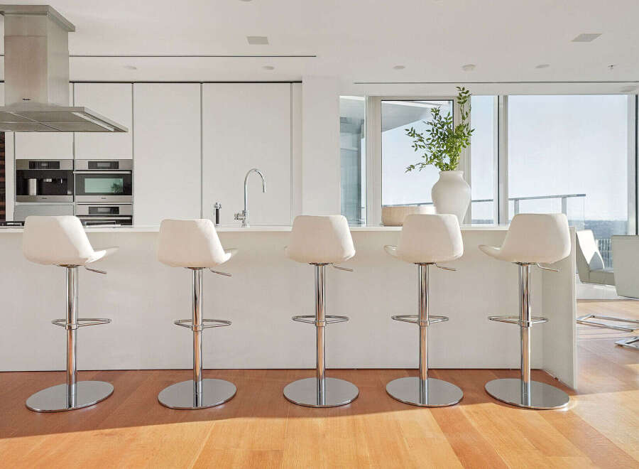 Modern kitchen with white cabinetry and stainless steel appliances, featuring a breakfast bar with four sleek white bar stools.
