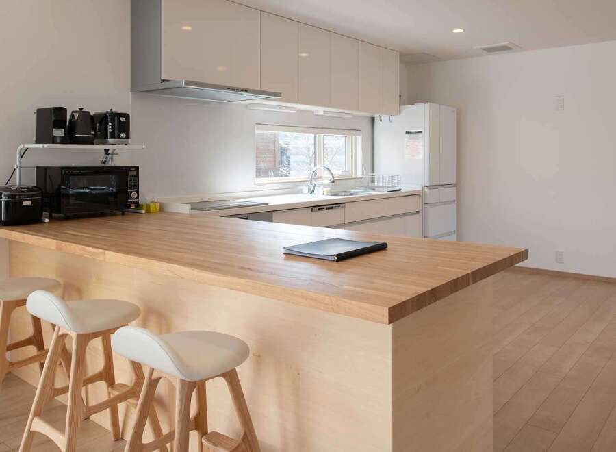 Modern kitchen with light wood countertops and white cabinets, featuring a breakfast bar with three minimalist stools.