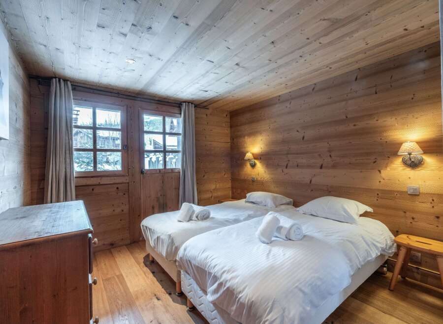 Cozy chalet bedroom with twin beds, wooden walls and ceiling, and a view of the snowy landscape.