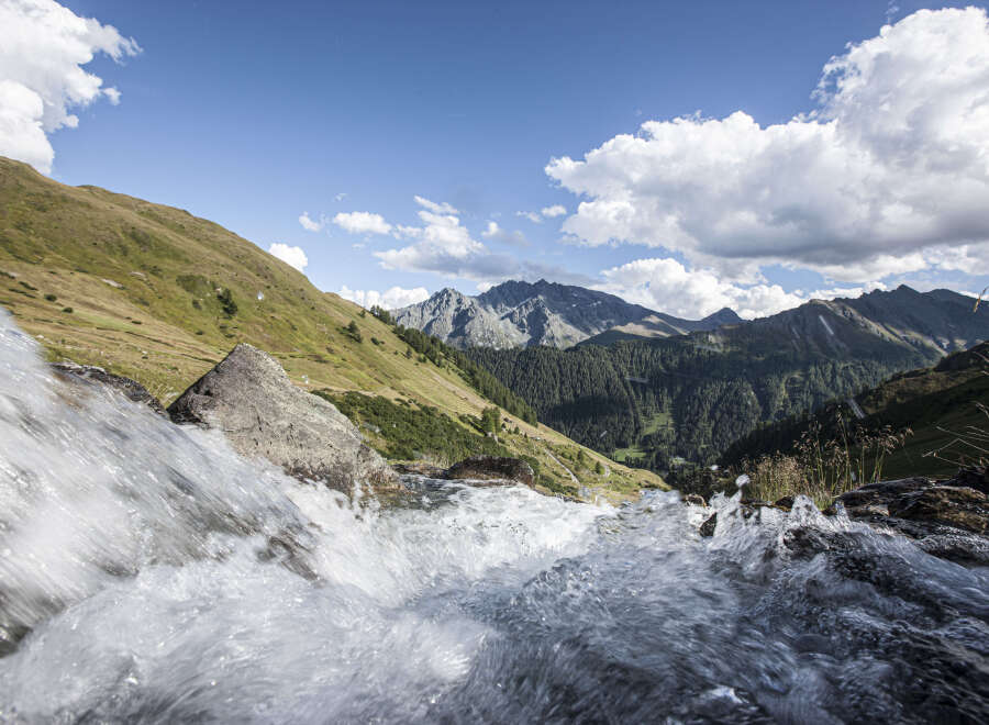 A cascading waterfall tumbles over rocks in the foreground, leading the eye to a breathtaking vista of rolling green hills and majestic mountains under a vibrant blue sky.