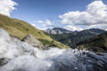 A cascading waterfall tumbles over rocks in the foreground, leading the eye to a breathtaking vista of rolling green hills and majestic mountains under a vibrant blue sky.