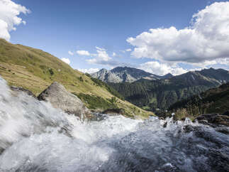 A cascading waterfall tumbles over rocks in the foreground, leading the eye to a breathtaking vista of rolling green hills and majestic mountains under a vibrant blue sky.