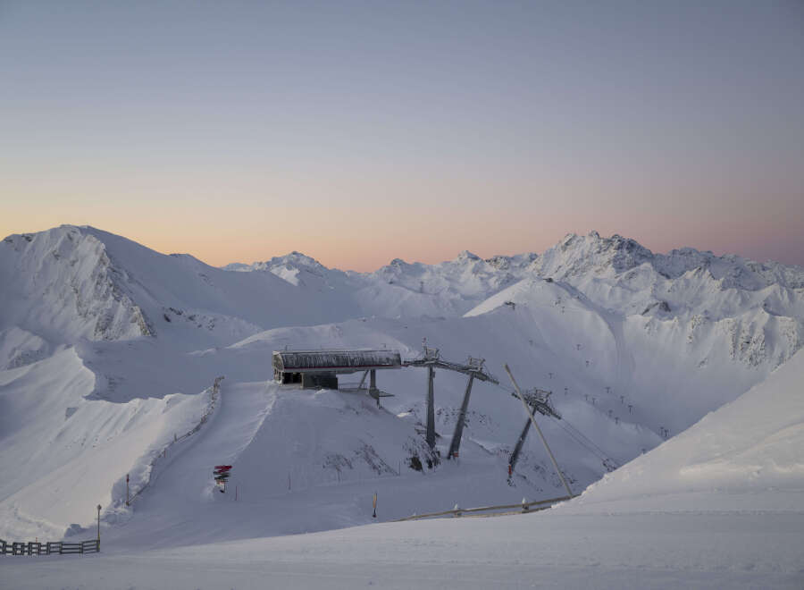Sunrise paints the snow-capped Austrian Alps in soft pink hues, as seen from a ski lift station.