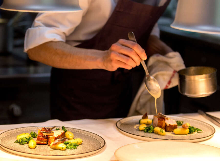 A chef carefully pours a creamy sauce over a plated dish of roasted meat and potatoes.