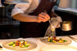 A chef carefully pours a creamy sauce over a plated dish of roasted meat and potatoes.