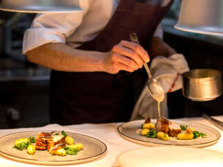 A chef carefully pours a creamy sauce over a plated dish of roasted meat and potatoes.