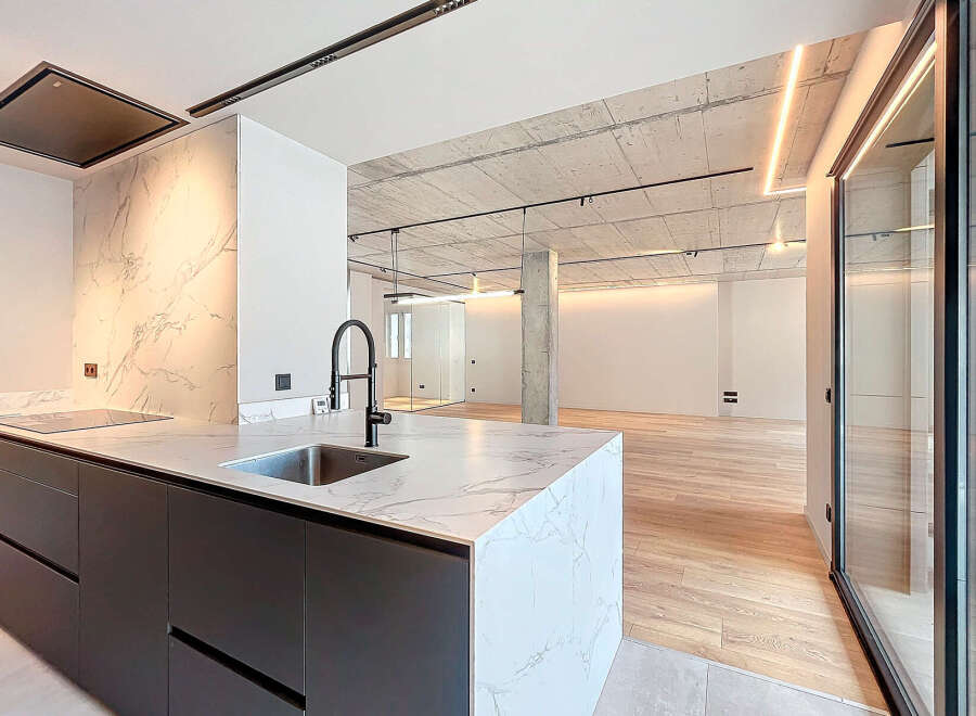 Modern kitchen with marble countertops and dark gray cabinetry, open to a spacious living area with hardwood floors and exposed concrete ceilings.