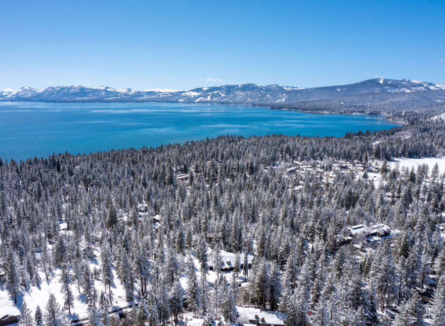 This stunning aerial view captures a snow-dusted forest with cozy cabins nestled amongst the evergreens, overlooking a crystal-clear lake and majestic snow-capped mountains in the distance.