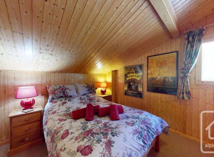Cosy chalet bedroom with floral bedding, wooden walls and ceiling, and a window with curtains.