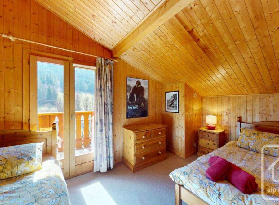 Cosy chalet bedroom with twin beds, wooden walls and ceilings, and a balcony offering stunning mountain views.