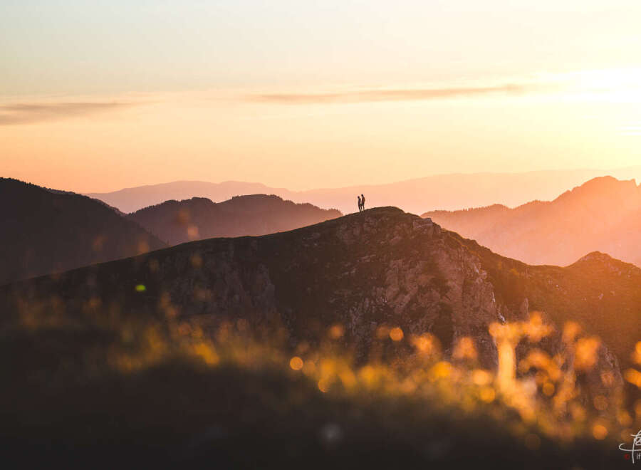 Silhouetted figures stand atop a majestic mountain peak, bathed in the golden light of a breathtaking sunset.