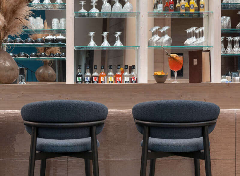 Modern bar with sleek, dark-colored bar stools and a back wall of glass shelving displaying an array of wine and liquor bottles.