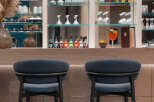 Modern bar with sleek, dark-colored bar stools and a back wall of glass shelving displaying an array of wine and liquor bottles.