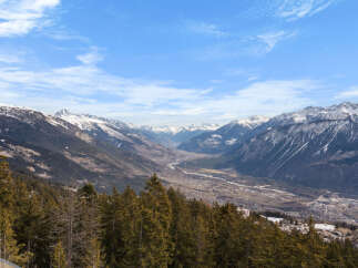 Breathtaking panoramic view of a snow-capped mountain range and valley, showcasing a picturesque landscape with a dense forest in the foreground.