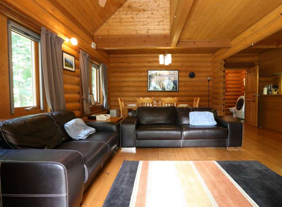 Cozy log cabin living room with leather sofas, a wood dining table, and large windows offering scenic views.