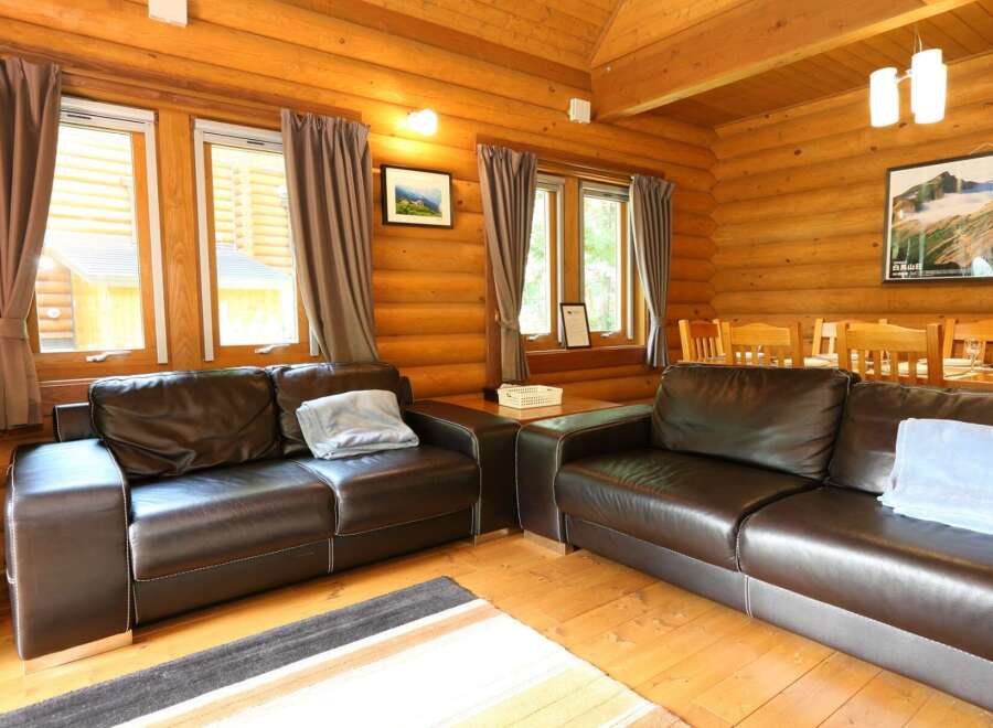 Cozy log cabin living room with two brown leather sofas, large windows with curtains, and a wooden dining table.