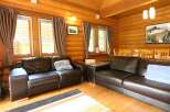Cozy log cabin living room with two brown leather sofas, large windows with curtains, and a wooden dining table.