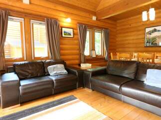 Cozy log cabin living room with two brown leather sofas, large windows with curtains, and a wooden dining table.