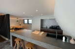 Modern kitchen with concrete countertops and wooden stools, open to a dining area with wooden table and chairs.